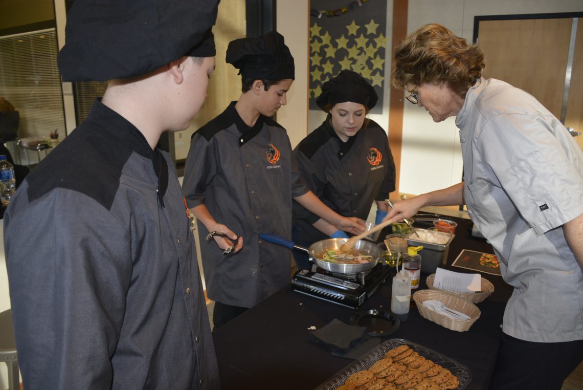 Brenda and three students in front of a table where student is preparing food as part of their culinary final