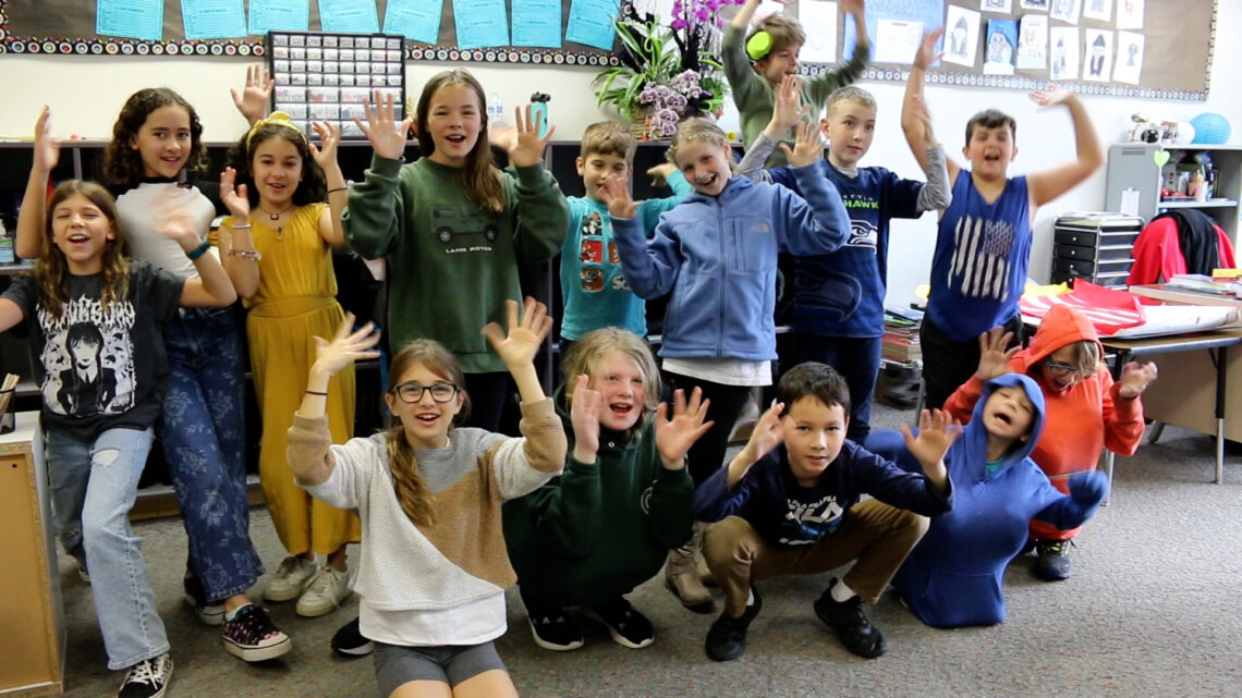 Group of 5th grade students wave their hands in the air energetically while smiling at the camera.